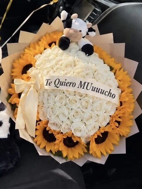 Sunflowers and pale blooms arranged around a plush cow, with a ribbon across the center.