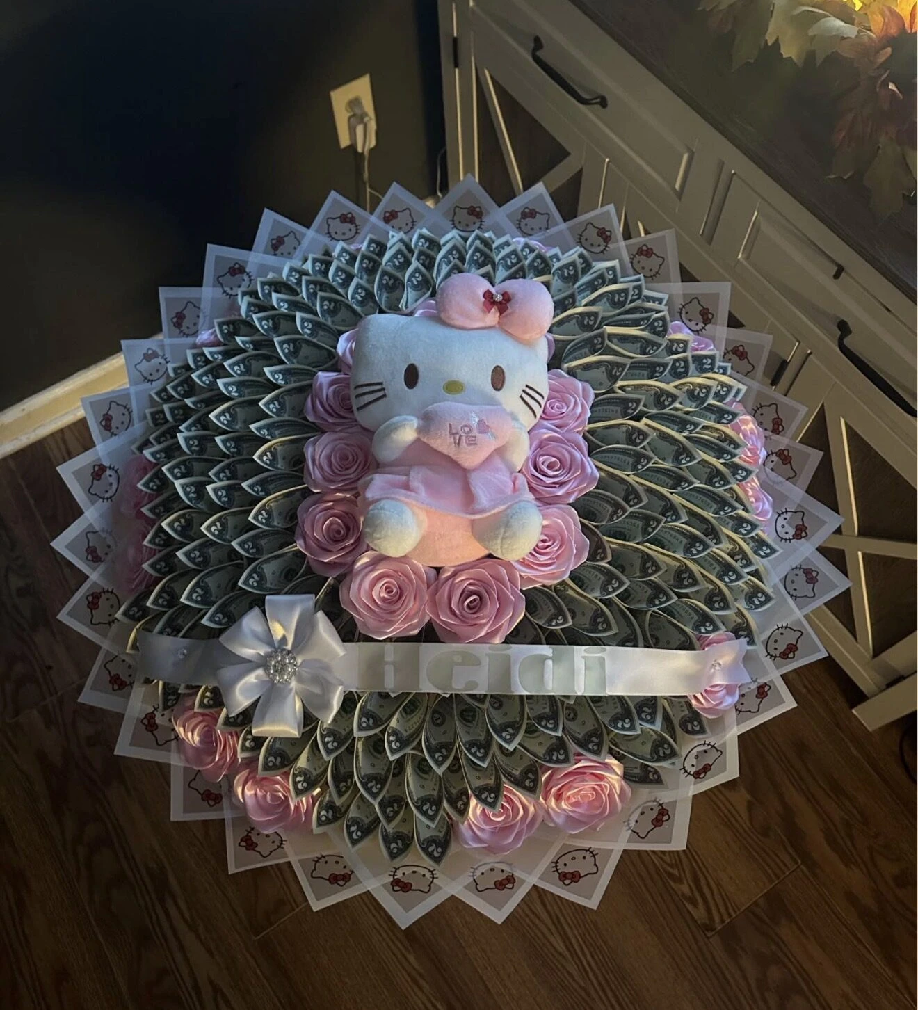 A round arrangement of folded bills with pink blooms and a small plush character at the center.