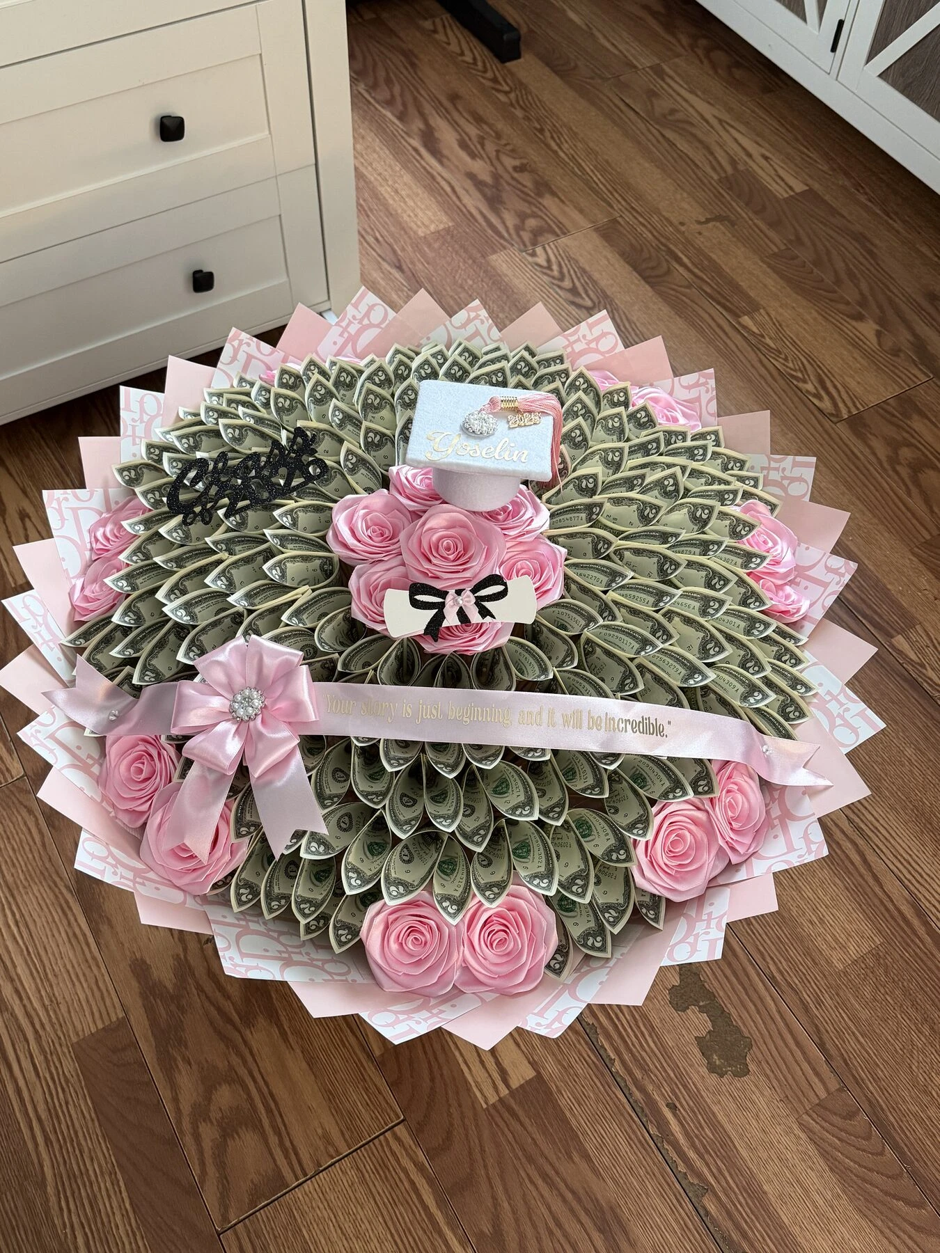 A round arrangement of folded bills with pink blooms and a pink bow.