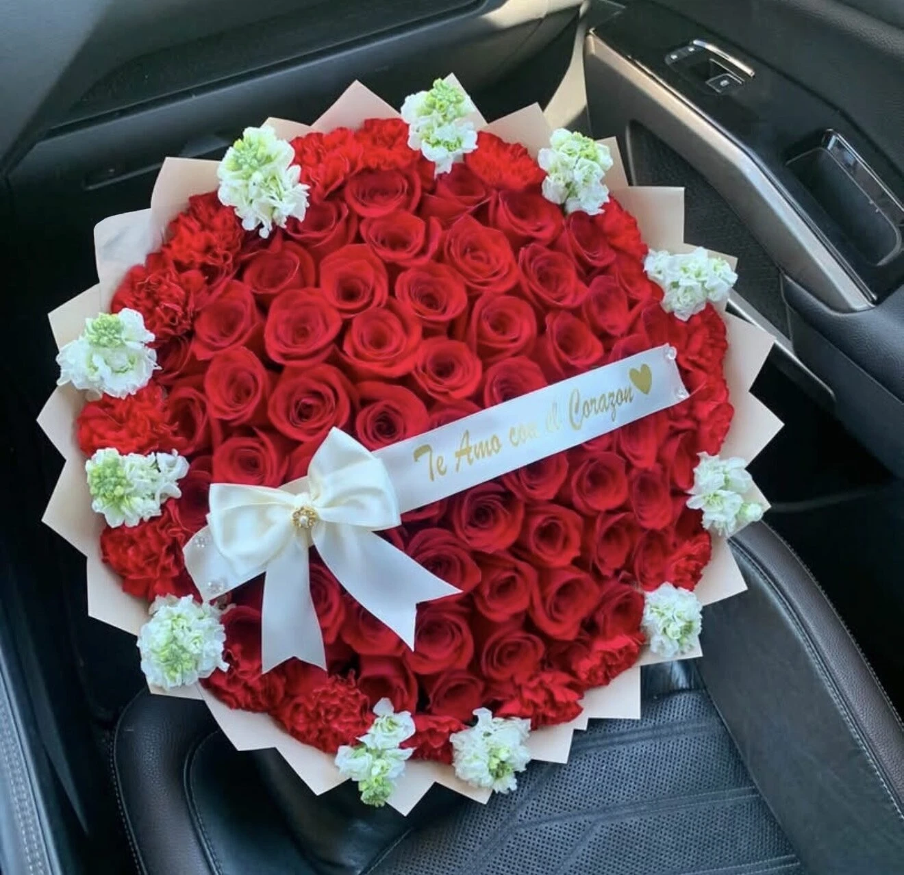A round red floral arrangement with white blooms and a pale bow, photographed in a car.